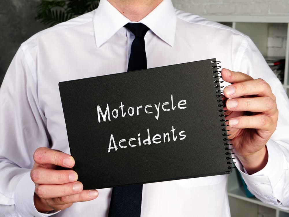A man in a white shirt holds a black notebook displaying the words "Motorcycle Accidents" in white chalk-like writing.