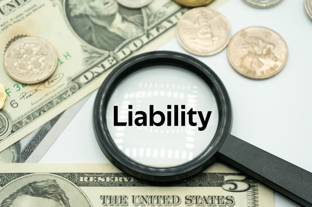 A magnifying glass prominently displays the word "Liability", surrounded by an array of U.S. paper bills and shiny coins.