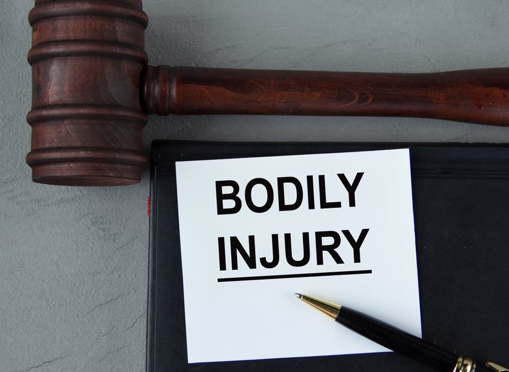 A wooden gavel beside a "BODILY INJURY" sign with a black pen on a gray surface.